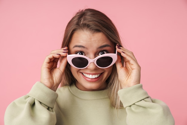 Portrait of beautiful smiling woman holding sunglasses and looking at camera isolated over pink wall