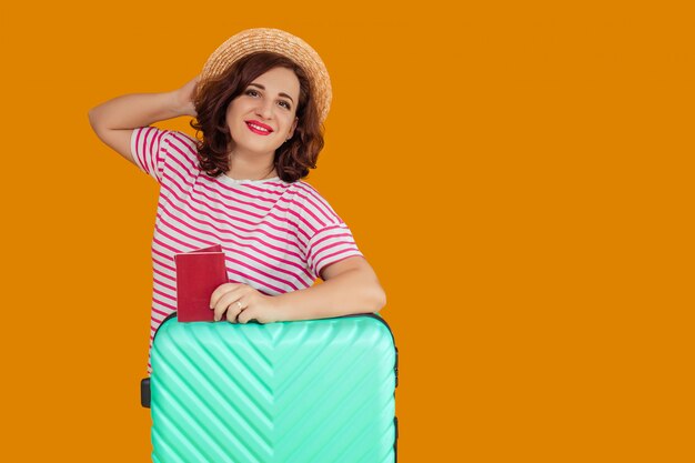 Portrait of a beautiful smiling woman with dark curly hair in a boater hat . a woman holds a passport in her hands and leans on a suitcase. awaiting travel