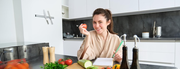 Portrait of beautiful smiling woman writing her healthy menu eating tomato while cooking making