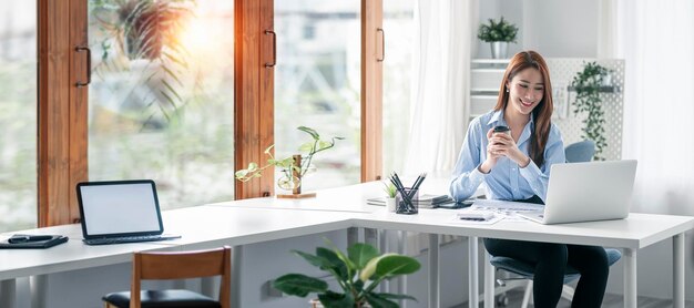 Portrait of beautiful smiling young entrepreneur businesswoman holding cup of coffee and working on laptop computer at office Panoramic background