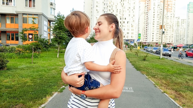 Portrait of beautiful smiling young mother holding her toddler son and walking on street