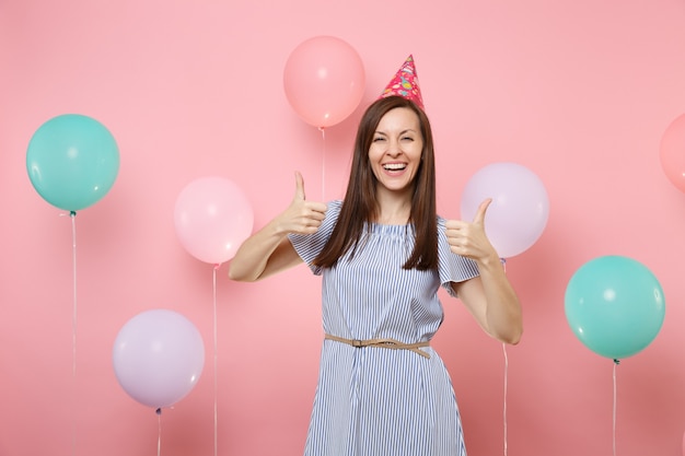 Portrait of beautiful smiling young woman in birthday hat and blue dress showing thumbs up on bright trending pink background with colorful air baloons. Birthday holiday party, people sincere emotion.