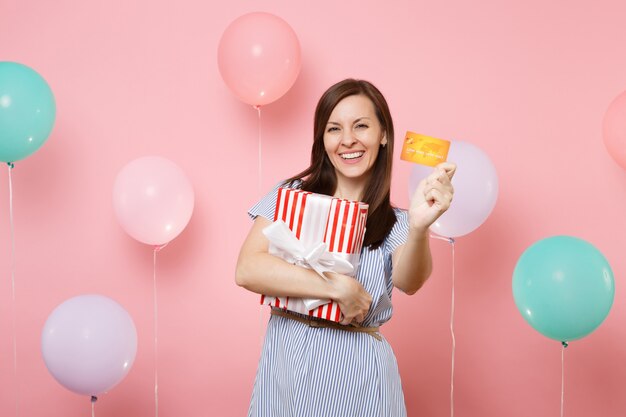 Portrait of beautiful smiling young woman in blue dress holding credit card and red box with gift present on pink background with colorful air balloons. Birthday holiday party, people sincere emotion.