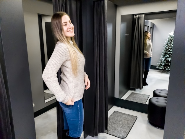 Portrait of beautiful smiling young woman trying on warm wool sweater in shopping mall dressing room