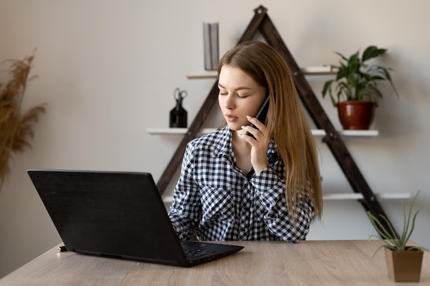 Portrait of a beautiful student who is talking on a cell phone and sitting at a table with a laptop open for work Gadgets the Internet and the mobile network in the work and study of teachers