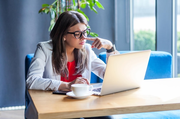 Portrait of beautiful stylish brunette young woman in glasses sitting looking at her laptop screen on video call and pointing at her nose indoor studio shot cafe office background