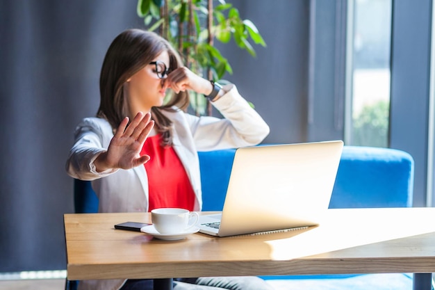 Portrait of beautiful stylish brunette young woman in glasses sitting pinching her nose and showing stop gesture because bad smell indoor studio shot cafe office background