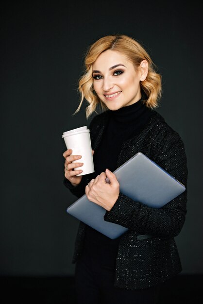 Portrait of a beautiful stylish businesswoman with a laptop and a glass of coffee in her hands and an open friendly smile. The concept of a strong independent woman.