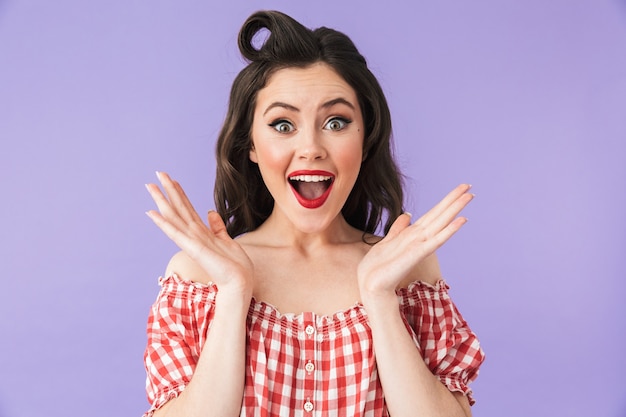 Portrait of a beautiful surprised pin-up girl wearing bright makeup standing isolated over violet wall, celebrating