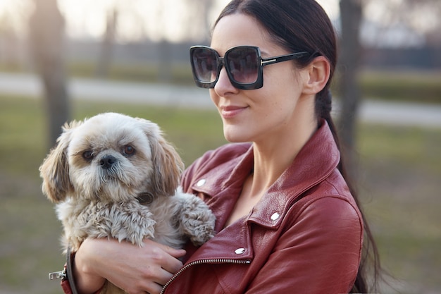 Портрет красивой сладкой милой женщины носить кожаную куртку и солнцезащитные очки, находясь на улице со своей собакой, проводить выходные с домашним животным, имея прогулку, стоя на улице.