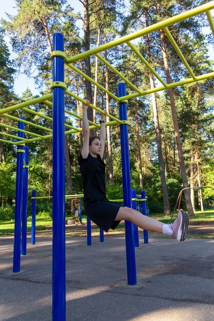 Portrait of beautiful teen age athletic girl training outdoors at open air gym at summer sunny day gain abdominal muscles on horizontal bar fitness healthy lifestyle