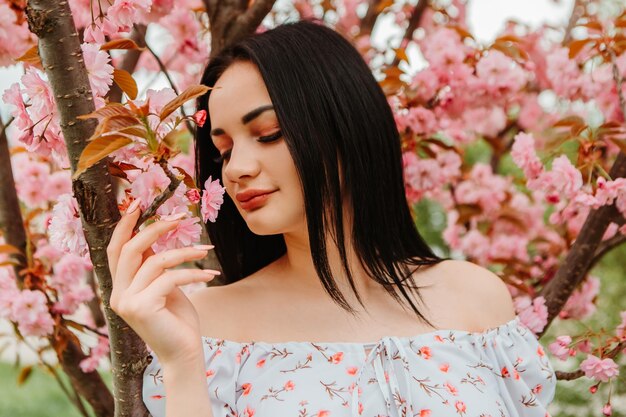 Portrait of beautiful tender woman dressed pink flowery dress posing near sakura cherry blossoms