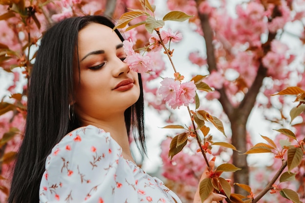 Portrait of beautiful tender woman dressed pink flowery dress posing near sakura cherry blossoms