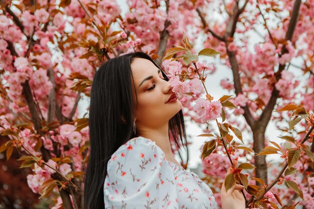 Portrait of beautiful tender woman dressed pink flowery dress posing near sakura cherry blossoms