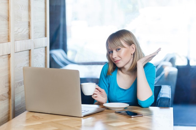 Portrait of beautiful thoughtful young girl with blonde bob haircut hair in blue blouse are sitting in cafe and talking with friend thoung webcame on laptop , looking to camera, drinking coffee.