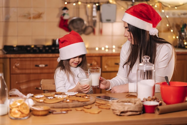 Portrait of beautiful tree year girl with her mom in santa hat sitting by the table in front of