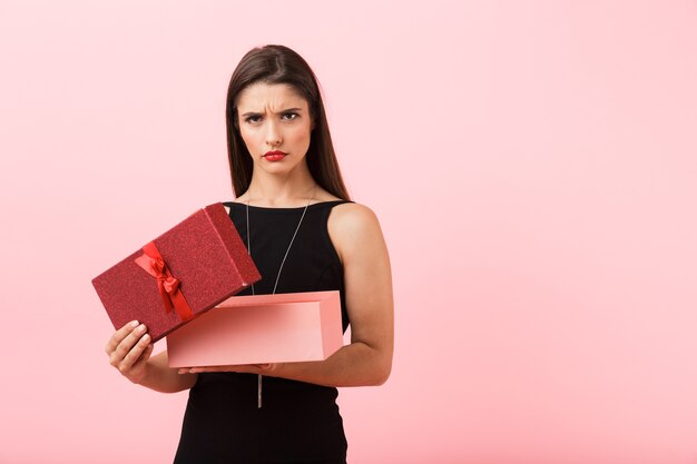 Portrait of a beautiful upset young woman wearing black dress standing isolated over pink background, open gift box