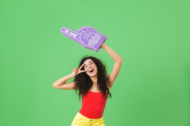 Portrait of beautiful woman 20s rejoicing and holding number one fan glove while standing on green wall