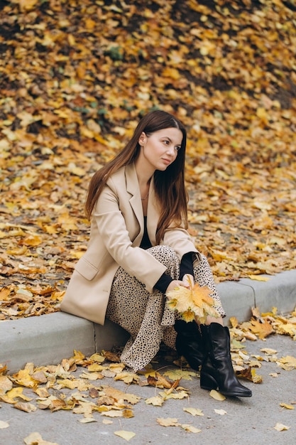 Ritratto di una bella donna in un cappotto beige che tiene foglie di acero gialle mentre si siede in un parco autunnale