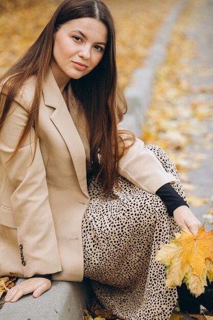 Portrait of a beautiful woman in a beige coat holding yellow maple leaves while sitting in an autumn park