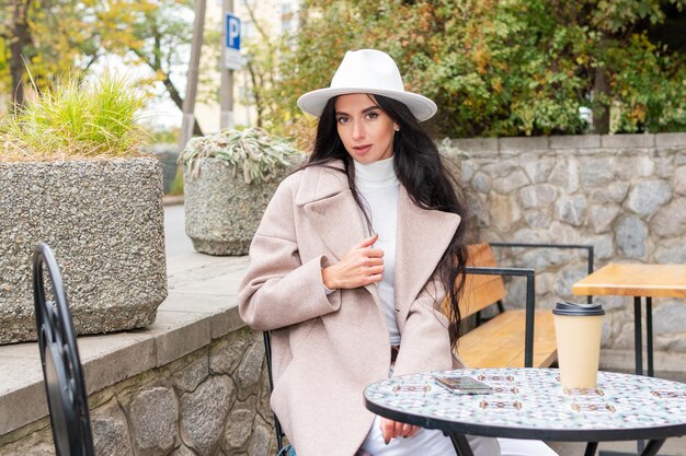 Portrait of a beautiful woman in a coat and hat at a table in a cafe on the street