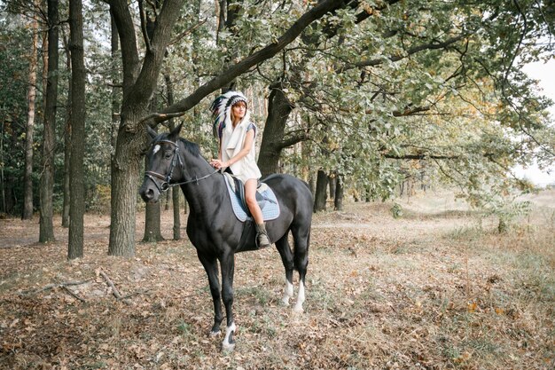 Портрет красивой женщины в платье и индийских перьях верхом на черной лошади на открытом воздухе Дневной снимок