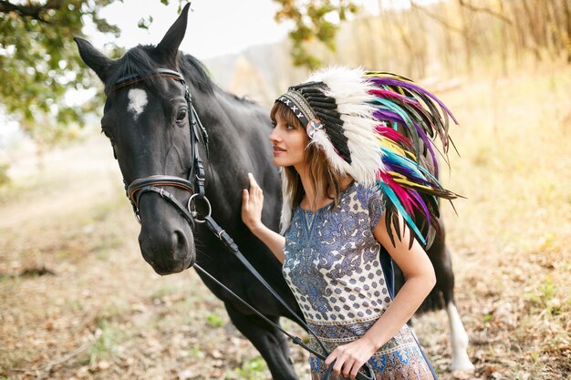 Portrait of a beautiful woman in a dress and indian feathers with a black horse outdoor Day shot