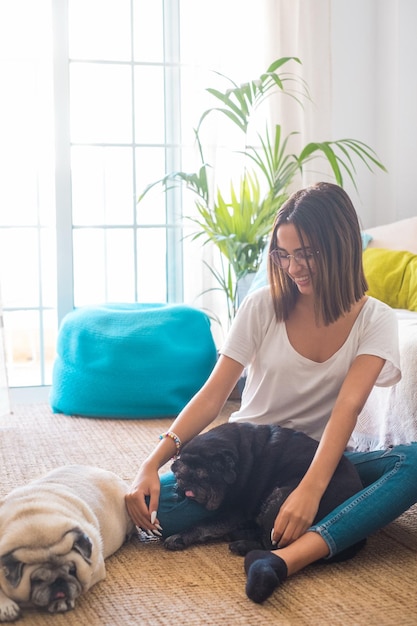 Portrait of beautiful woman in eyeglasses having fun with her pets pugs dogs sitting on floor in the living room of her house. Cheerful woman spending leisure time with her two cute pets dog at home