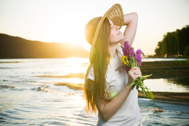 Portrait of a beautiful woman in a hat on a background of a sunset. Girl with a bouquet of flowers
