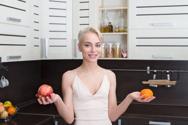 Портрет красивой женщины, держащей яблоко и апельсин на современной кухне