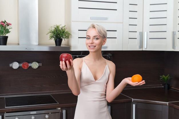 Portrait of beautiful woman holding apple and orange in modern kitchen