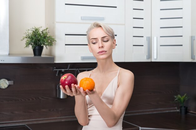 Portrait of beautiful woman holding apple and orange in modern kitchen