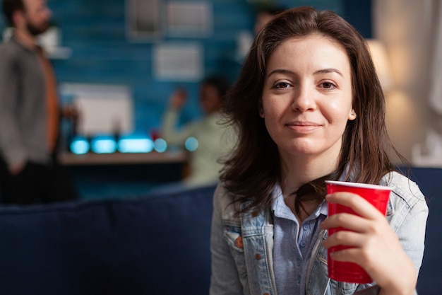 Photo portrait of beautiful woman holding beer glass looking into camera during night party. multi-ethnic diverse group of friends gathered together to celebrate birthday late at night cheerful young people