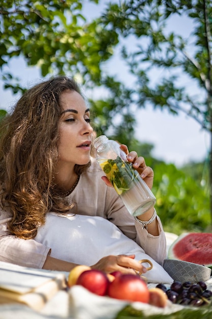 Ritratto di bella donna in posa sdraiato sul plaid al parco giardino estivo un libro frutta una borsa e un wat