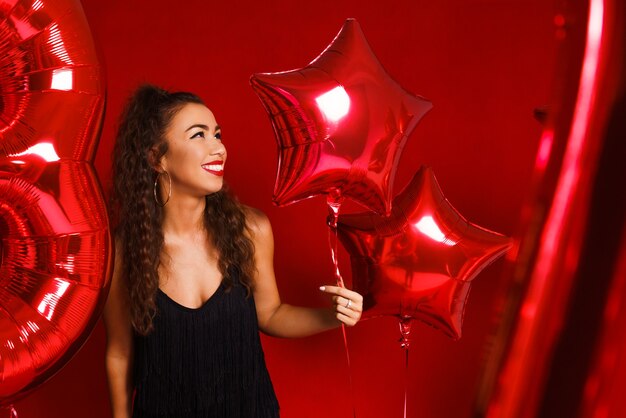 Portrait of a beautiful woman on a red background with red balloons posing in a black dress the girl...