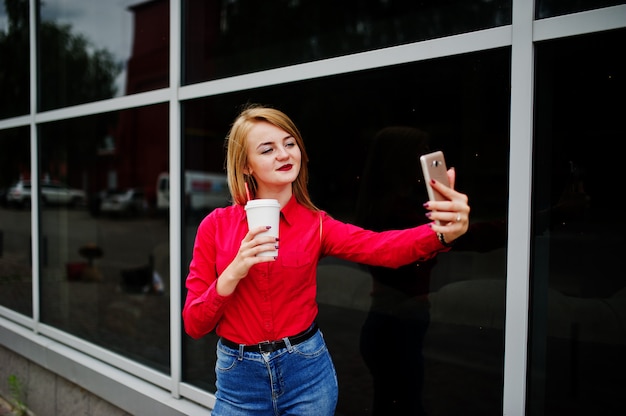 Portrait of a beautiful woman in red blouse and casual jeans taking selfie on mobile phone and holding a cup of coffee outside the huge shopping mall.