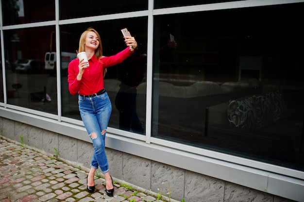 赤いブラウスと携帯電話でselfieを取り、巨大なショッピングモールの外のコーヒーカップを保持しているカジュアルなジーンズで美しい女性の肖像画。