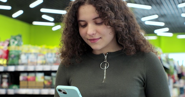 Portrait of beautiful woman use smartphone in supermarket Woman buyer using mobile phone application standing with shopping trolley in supermarket