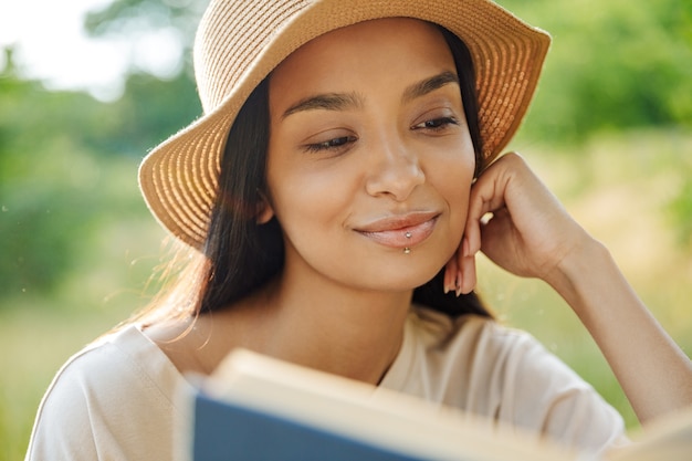 Портрет красивой женщины с пирсингом в губе и соломенной шляпе, читающей книгу, сидя на траве в зеленом парке
