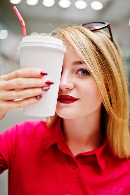 Portrait of a beautiful woman wearing red blouse, casual jeans and black high heels covers half of her face with a cup of coffee in huge shopping mall.