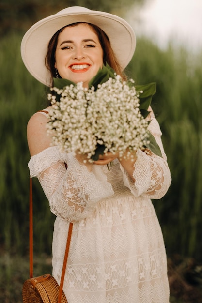 Portrait of a beautiful woman in a white dress and a hat with lilies of the valley at sunset A girl in nature Spring flowers