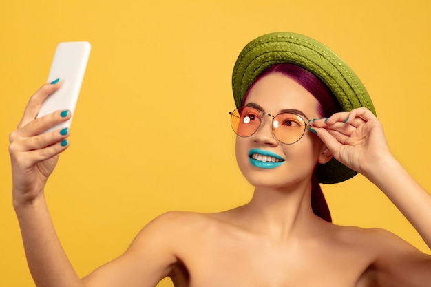 Portrait of beautiful woman with bright make-up, hat and sunglasses on yellow studio background. Stylish and fashionable make, hairstyle. Colors of summer. Beauty, fashion, ad concept. Takes selfie.