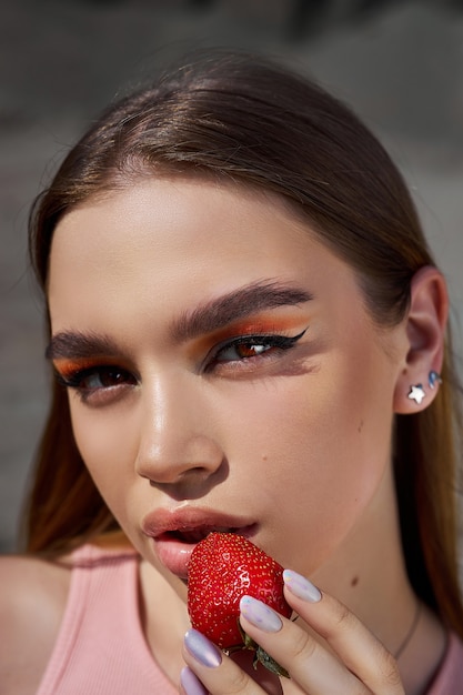 Ritratto di una bella donna con trucco rosso brillante e una fragola in primo piano mano. ombre rosse sugli occhi di una ragazza, trucco professionale, cosmetici naturali