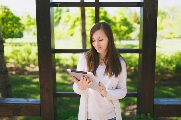 Portrait of beautiful woman with charming smile wearing light casual clothes. Pretty girl holding tablet pc computer, reading news in city park in street outdoors on spring nature. Lifestyle concept.