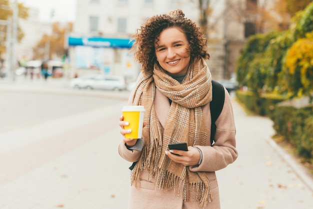 Portrait of beautiful woman with curly hair holding cup of coffee take away and looking at the camera