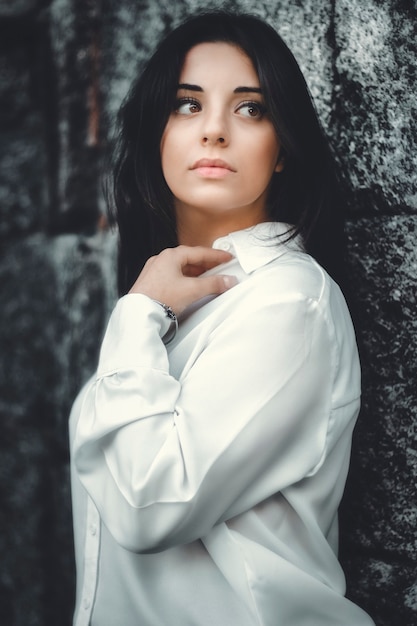 Portrait of a beautiful woman with dark hair in a white shirt posing on a stone wall