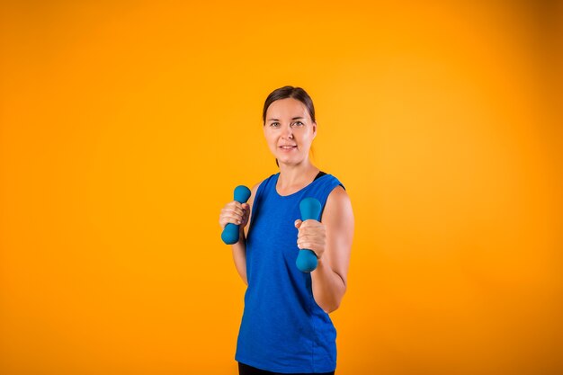 Portrait of a beautiful woman with dumbbells on an orange wall with a copy of space