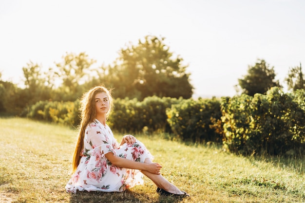 portrait of beautiful woman with long curly hair on currant field. Girl in a light dress sits on the grass in sunny day