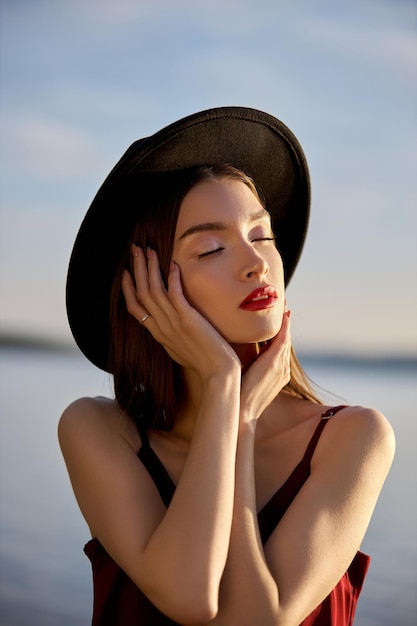 Portrait of a beautiful woman with long hair in a big round black hat. Beauty skin face, bare shoulders. Girl golden light at sunset