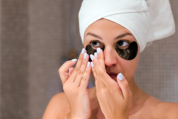 Portrait of beautiful woman wrapped in a towel is applying black hydrogel eye patches looking in the mirror Spa procedure in the morning